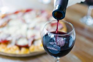 Red wine is being poured into a glass in front of a whole pizza